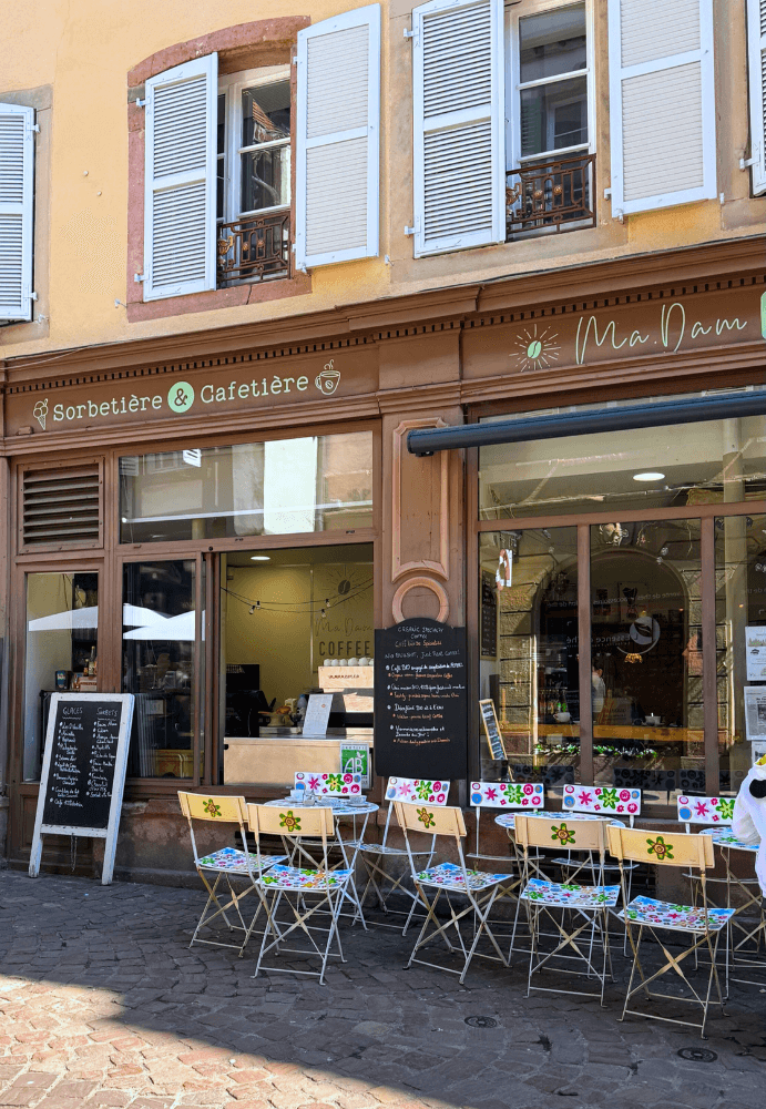 Devanture du glacier Ma.Dam Coffee à Colmar, avec terrasse colorée et ardoises affichant les parfums de glaces artisanales, situé dans une rue pittoresque du centre-ville